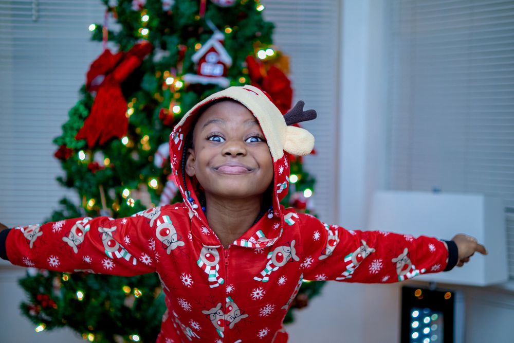 A person in a red holiday pajama set spreads their arms wide in front of a Christmas tree.