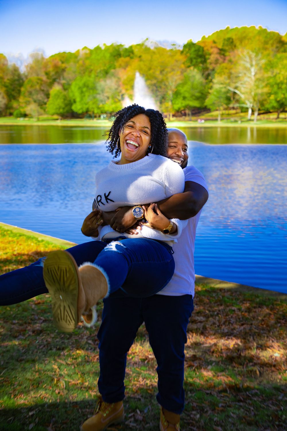 Playful moment captured by lake with fountain and autumn trees in background.