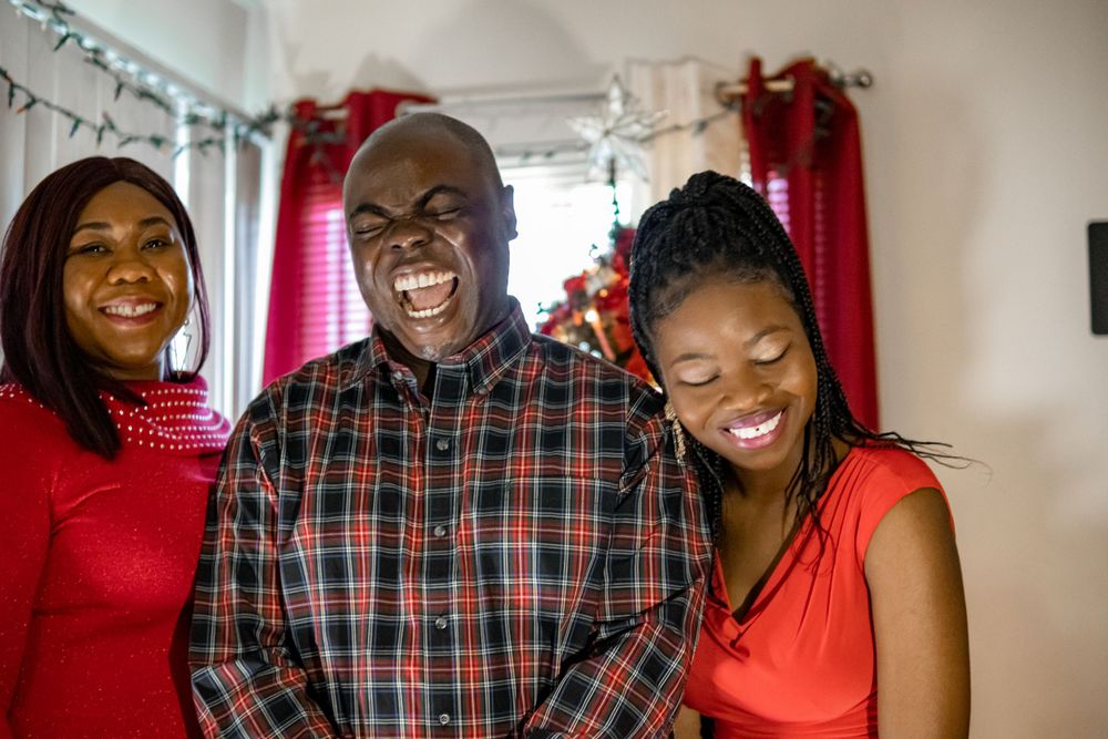 Three people smile and laugh together during Christmas celebration with red curtains.