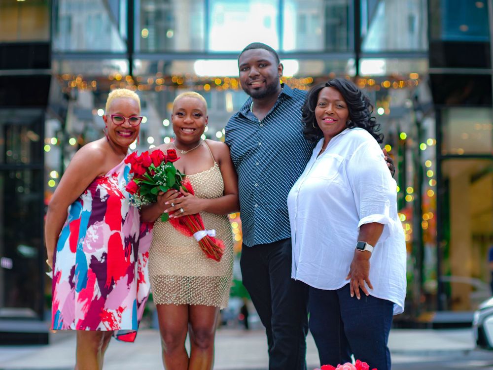 A small group in colorful attire poses together on an urban street with bouquets of red flowers.