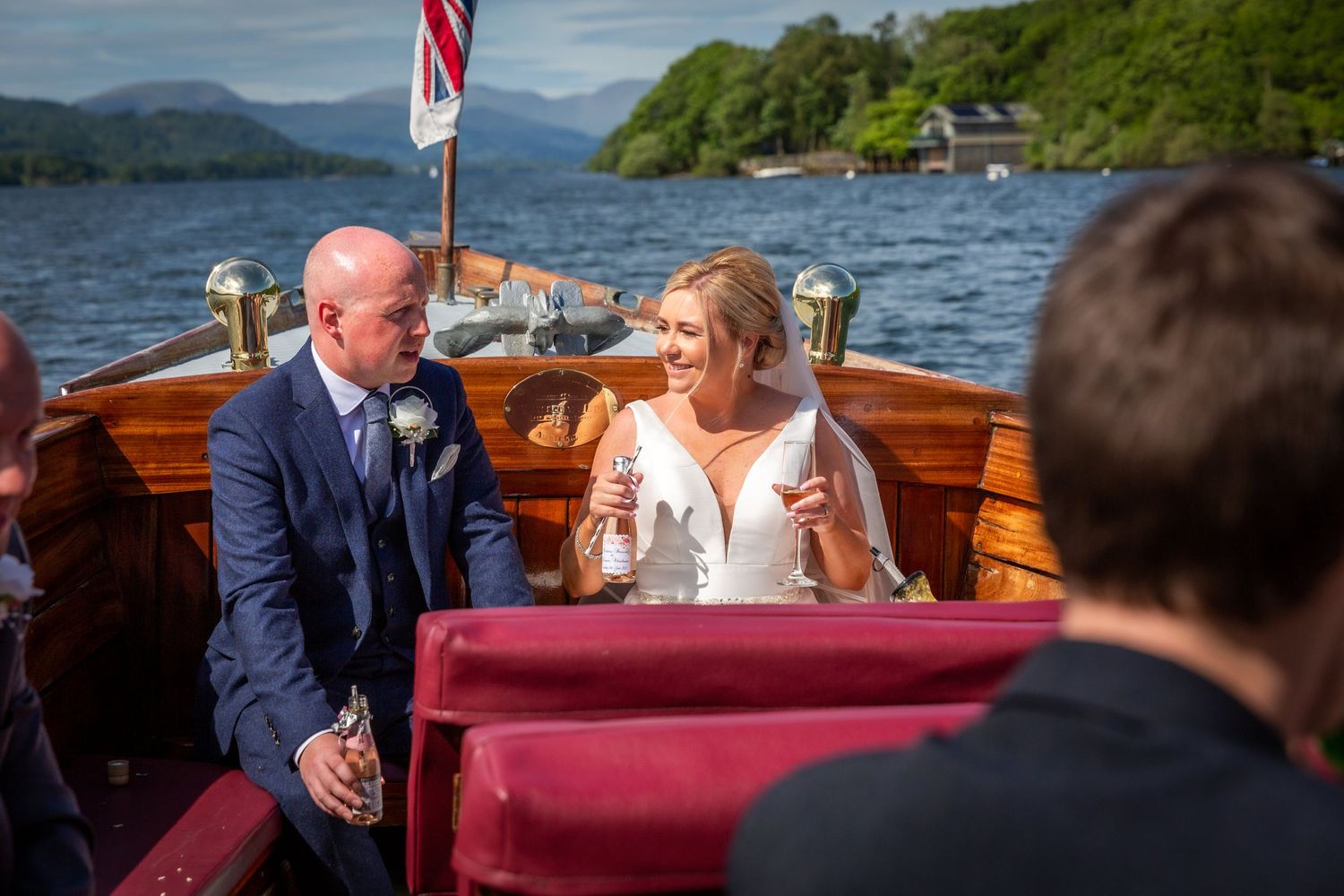 groom enjoys a beer on the wooden boat as the bride holds a glass of champagne in each hand, lakeland fells behind them