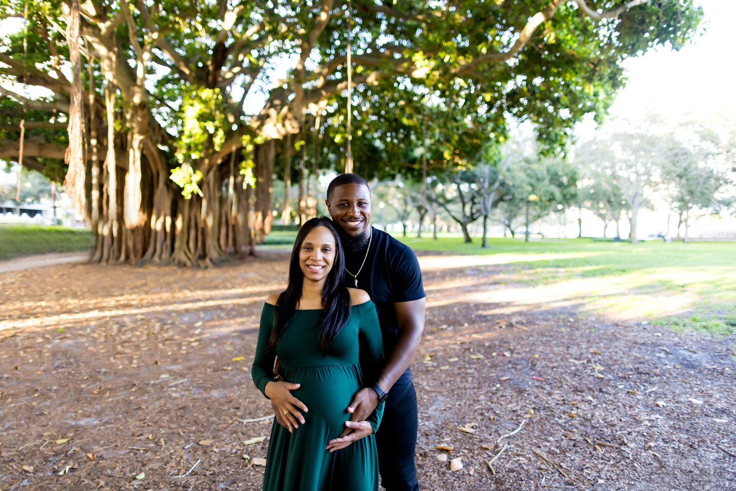 Maternity photo of couple under large banyan trees in park setting.