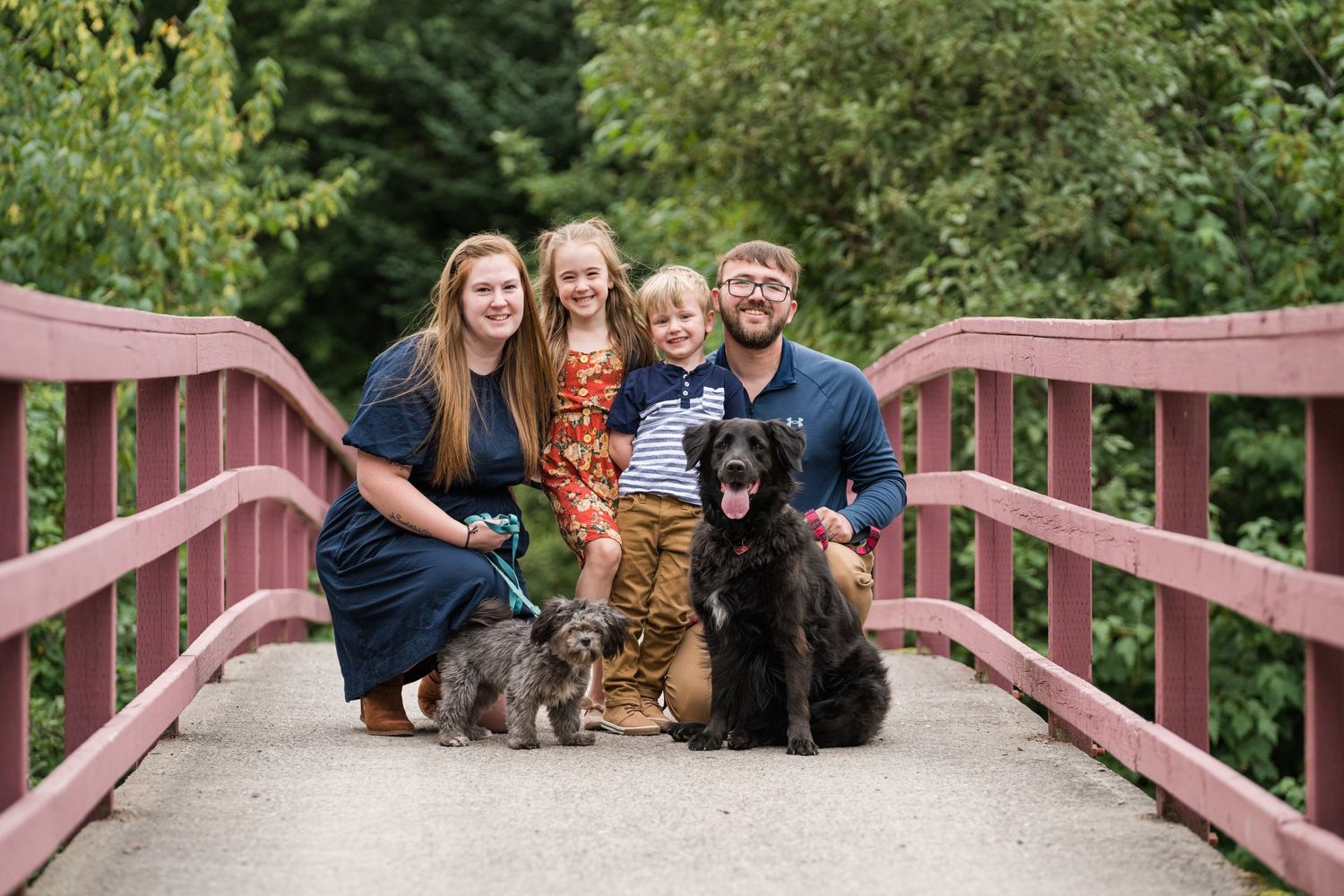 Family portrait with three dogs on red wooden bridge surrounded by green foliage.