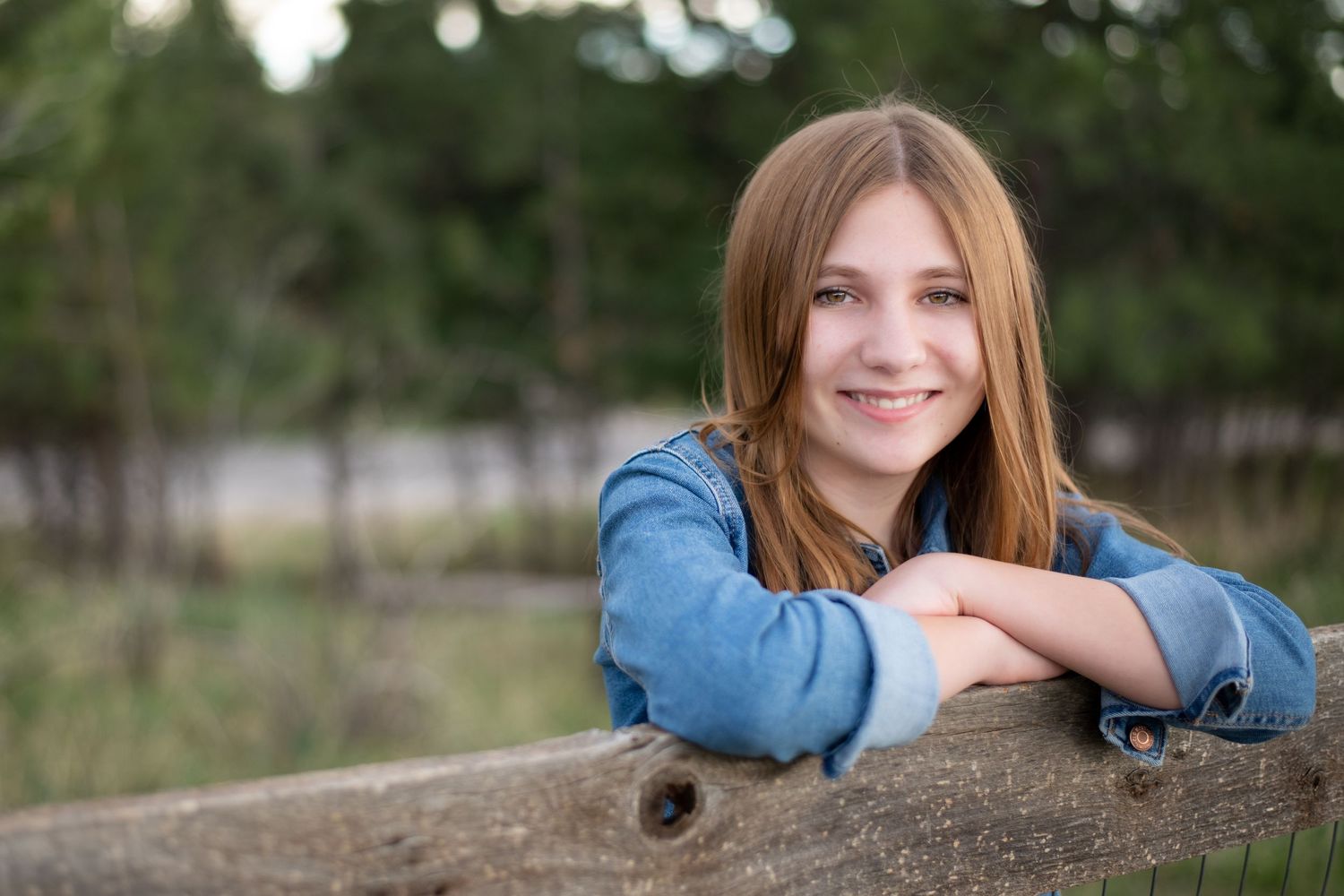 A portrait in nature shows a person in denim leaning on a wooden fence with a soft bokeh background.