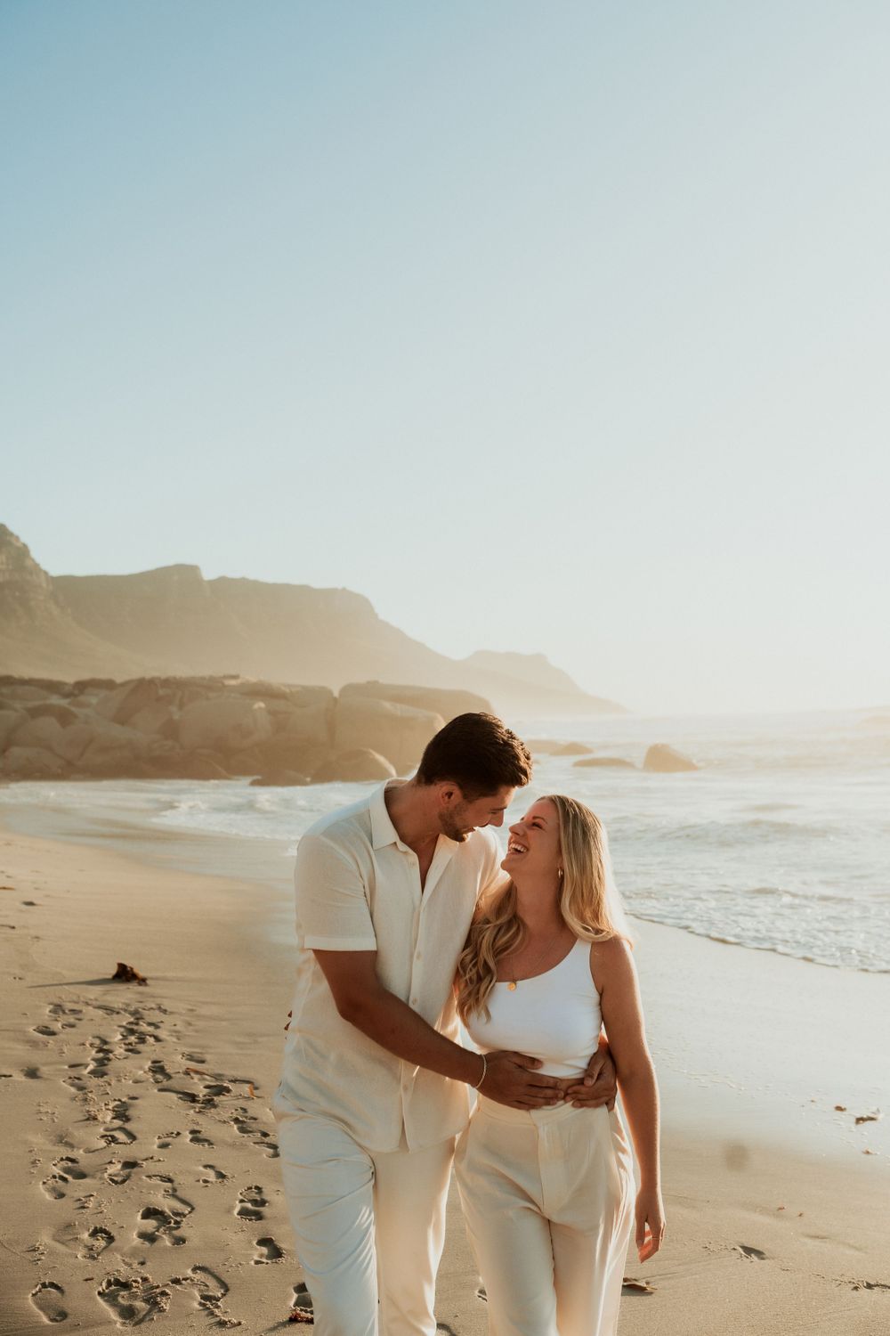 A couple in white outfits walks along a scenic beach at sunset.