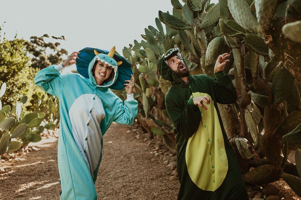 Two people in dinosaur costumes posing playfully next to cactus plants.
