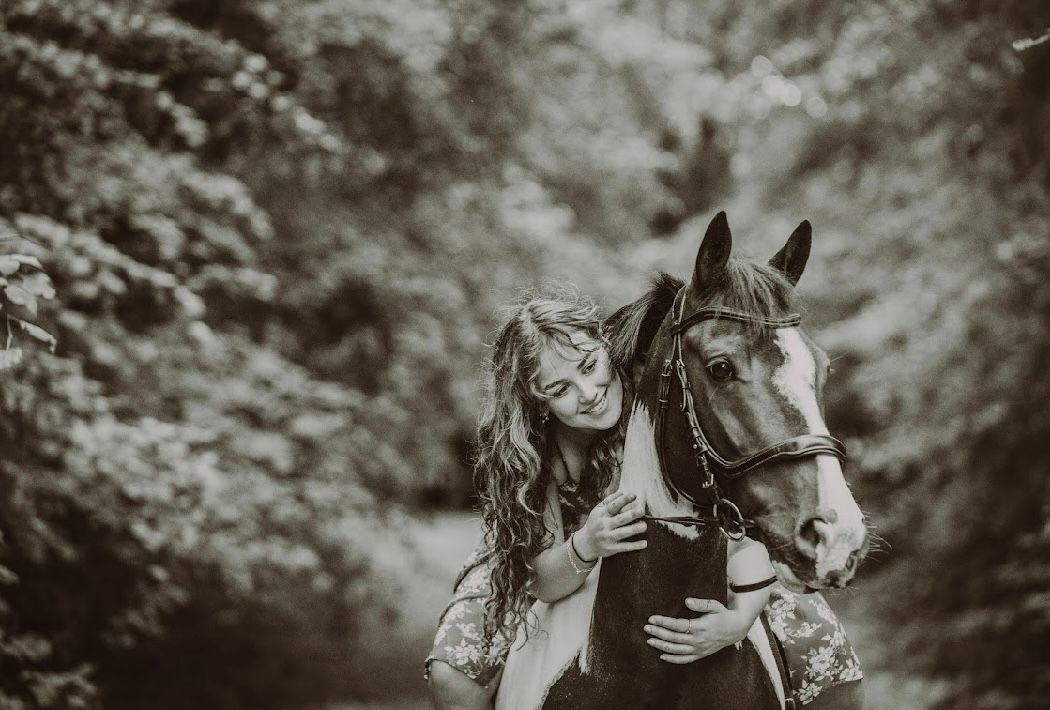 A young woman with curly hair smiling and embracing a brown horse in a lush green forest setting.