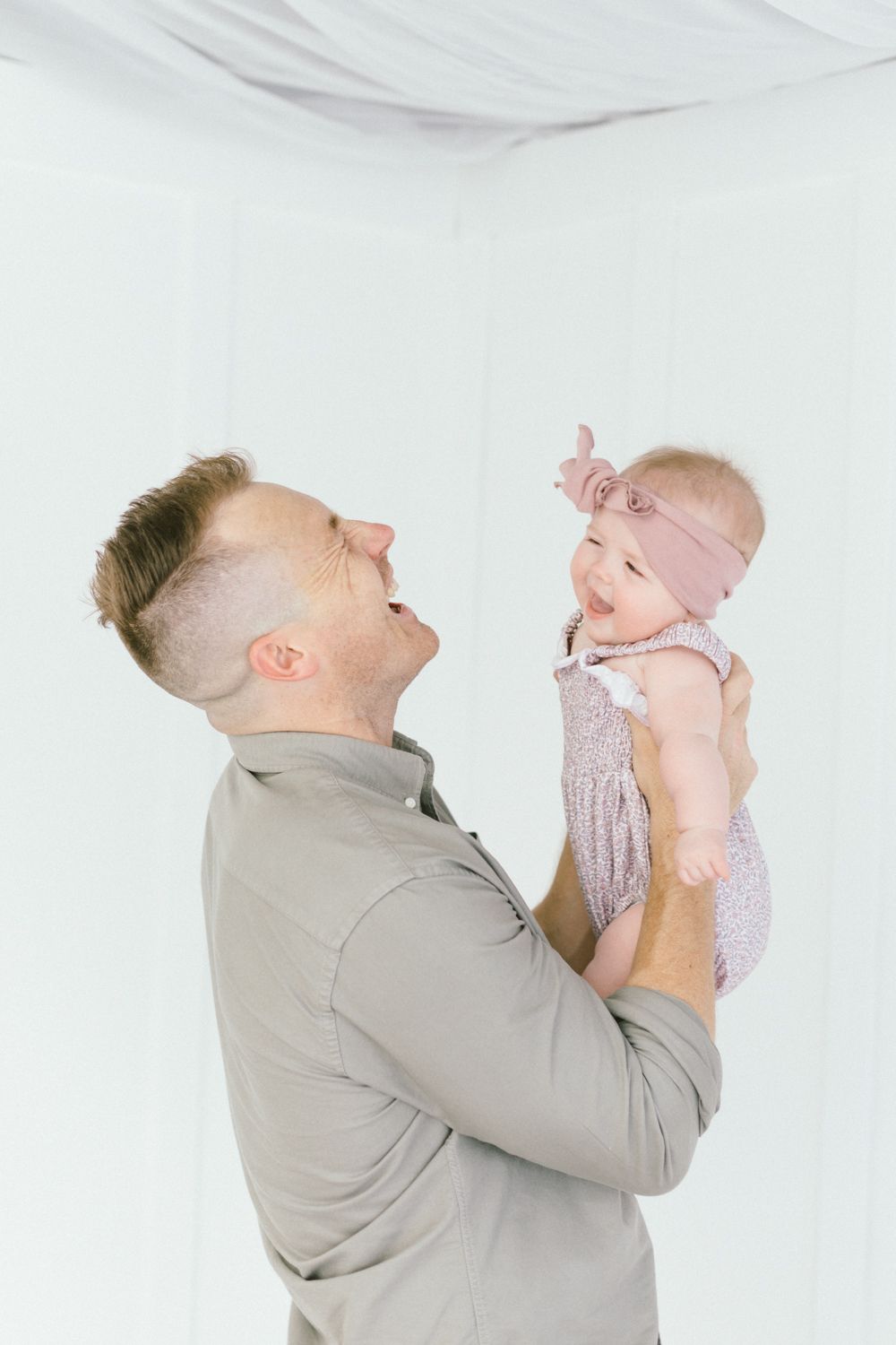 Parent lifting baby in pink dress with joyful expression against white background.