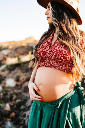 A close-up maternity portrait with sun hat and red floral top against rocky landscape.