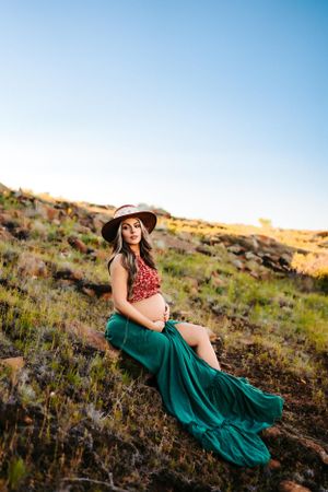 A maternity portrait at sunset featuring a woman in emerald skirt and red top on rocky terrain.