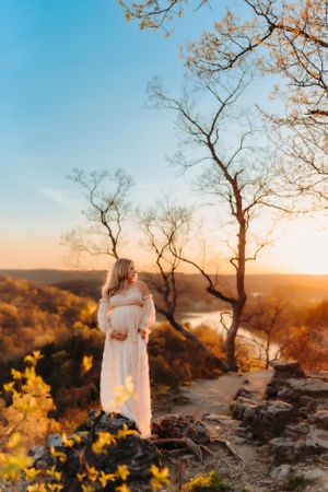 A person in a flowing white dress stands on rocky terrain during sunset.