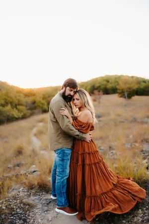 Romantic outdoor portrait with flowing rust dress against golden hillside at sunset.
