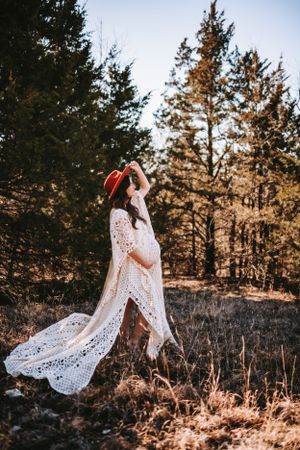 An ethereal maternity portrait features a flowing lace dress against evergreen trees.