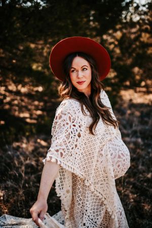 A boho maternity portrait features a red hat and delicate lace dress in golden sunlight.