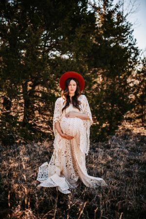 A peaceful maternity portrait showcases an elegant white lace gown and red hat in nature.