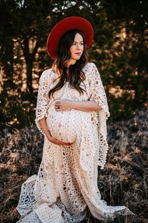 A serene maternity portrait shows a flowing lace dress and red hat against pine trees.