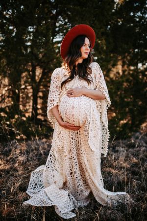 Expectant mother in bohemian lace dress and red hat poses at sunset in nature.