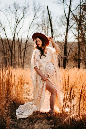 Romantic maternity portrait with red hat and lace dress in sunlit meadow.