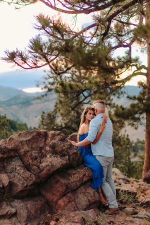 Two people share an intimate moment on a mountain cliff with evergreen trees and scenic views.