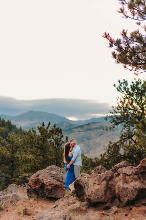 A couple embraces on a rocky mountain overlook with pine trees and mountain vista in the background.