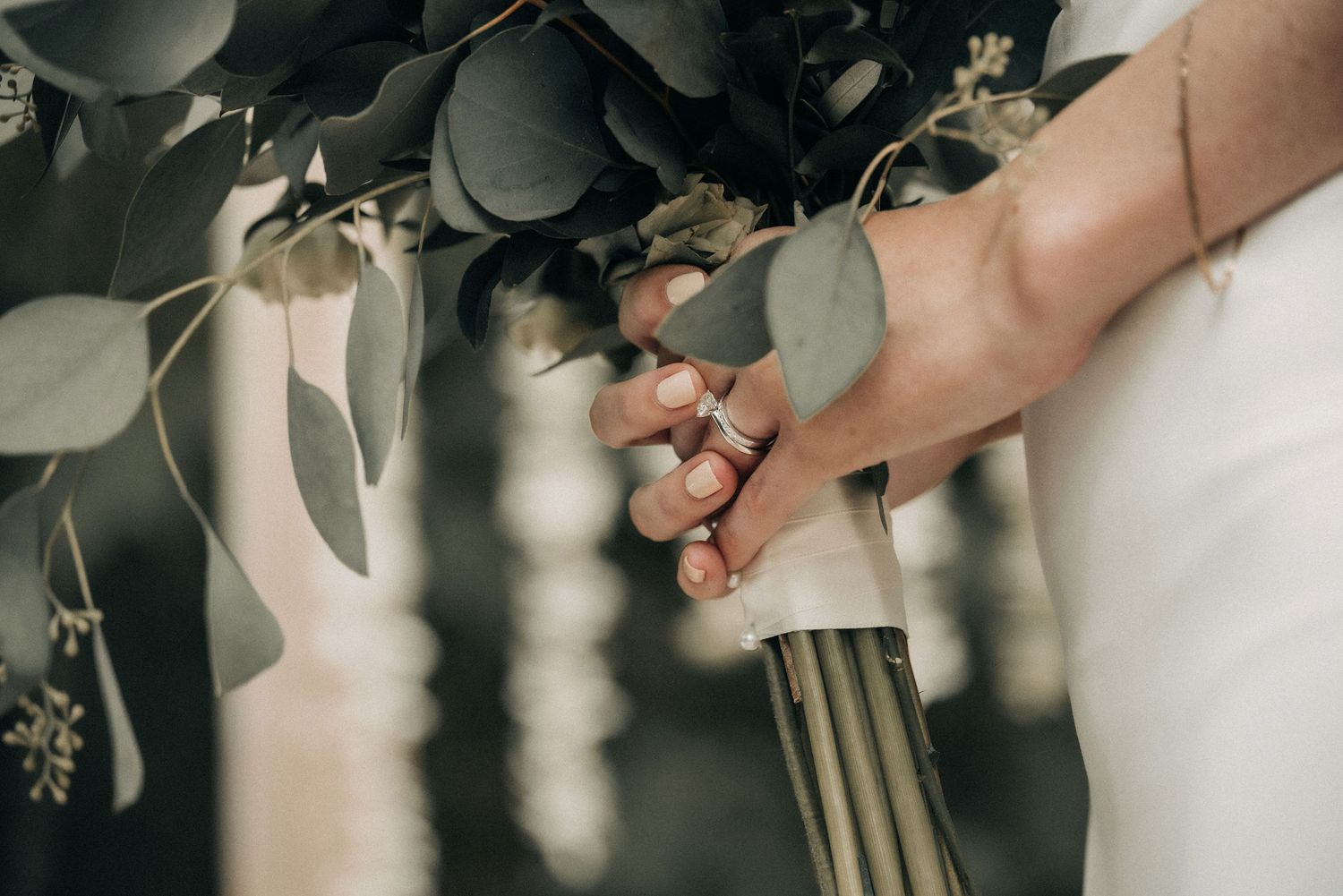 Delicate hands holding a wedding bouquet wrapped in white ribbon against dark foliage.