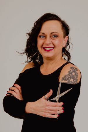 Woman in black top poses playfully with scissors against gray background.