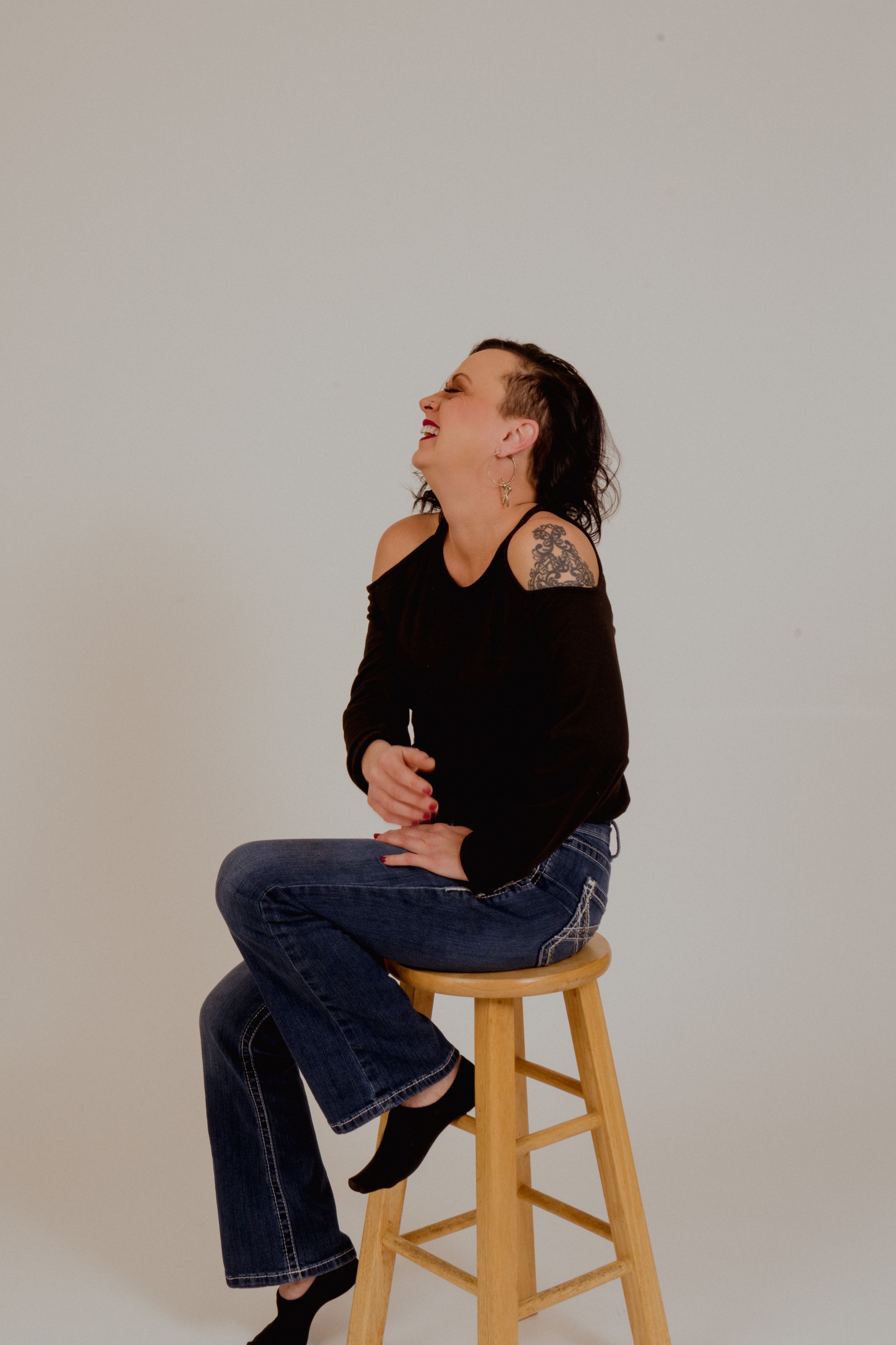 Person laughs while sitting on wooden stool in casual studio photoshoot.