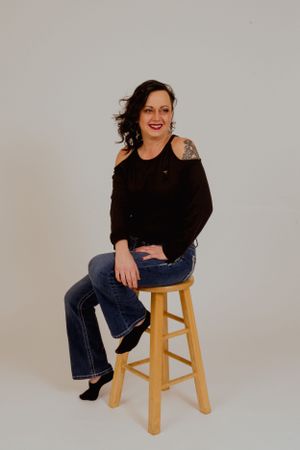 Person in black top and jeans poses expressively on wooden stool.