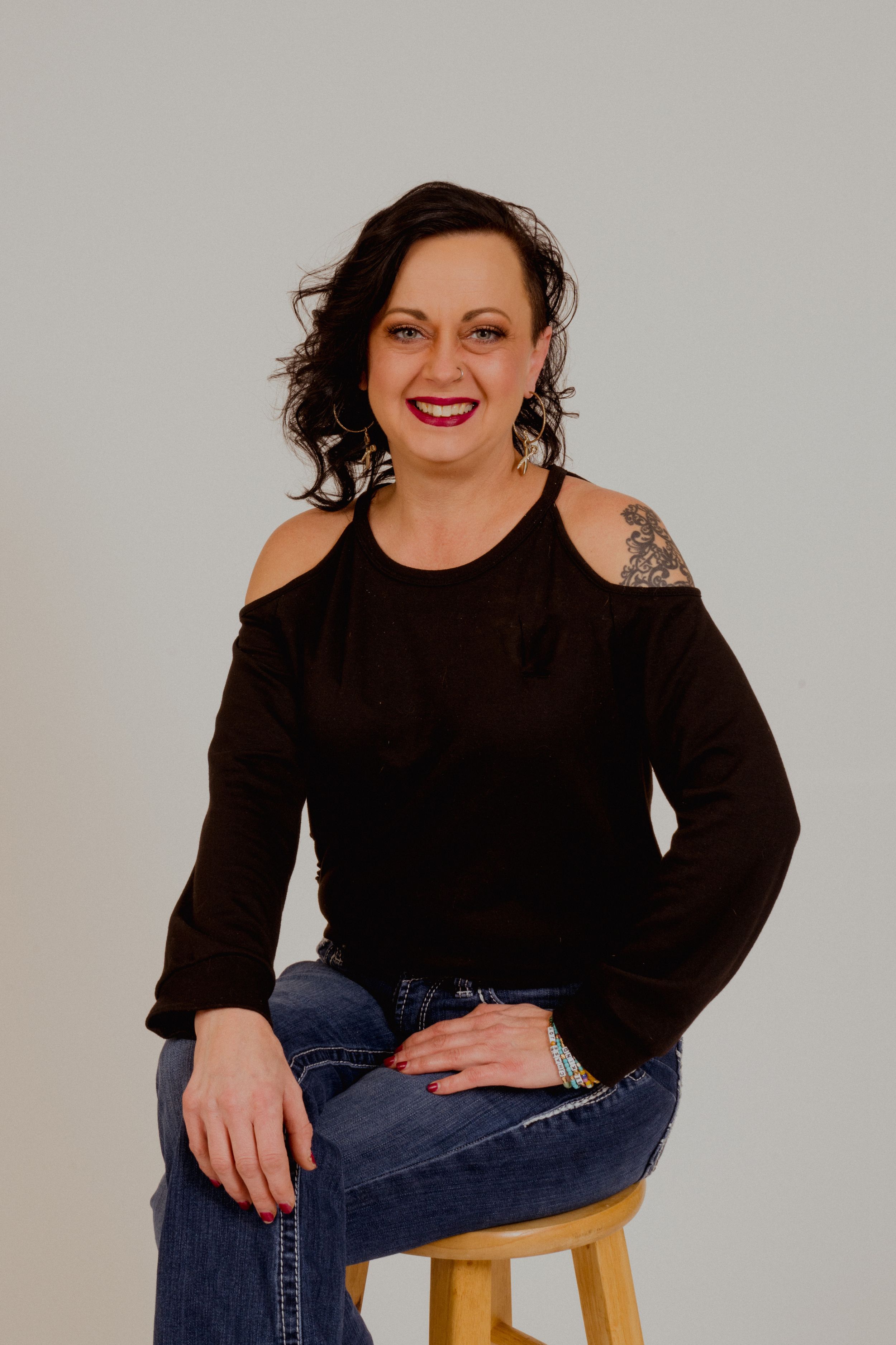 Person in black cold-shoulder top and jeans seated casually on wooden stool.