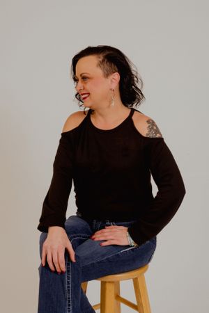 Casual portrait on wooden stool showing relaxed pose in black top and denim.