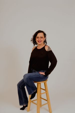 Person in black top and jeans sits casually on wooden stool against light background.