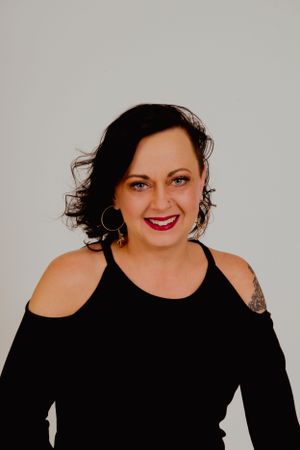 Person in black top with cold shoulders smiles while posing against neutral backdrop.