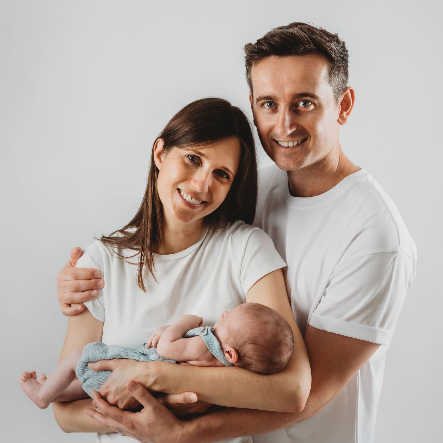 A tender family portrait with a newborn baby in light blue against a gray background.