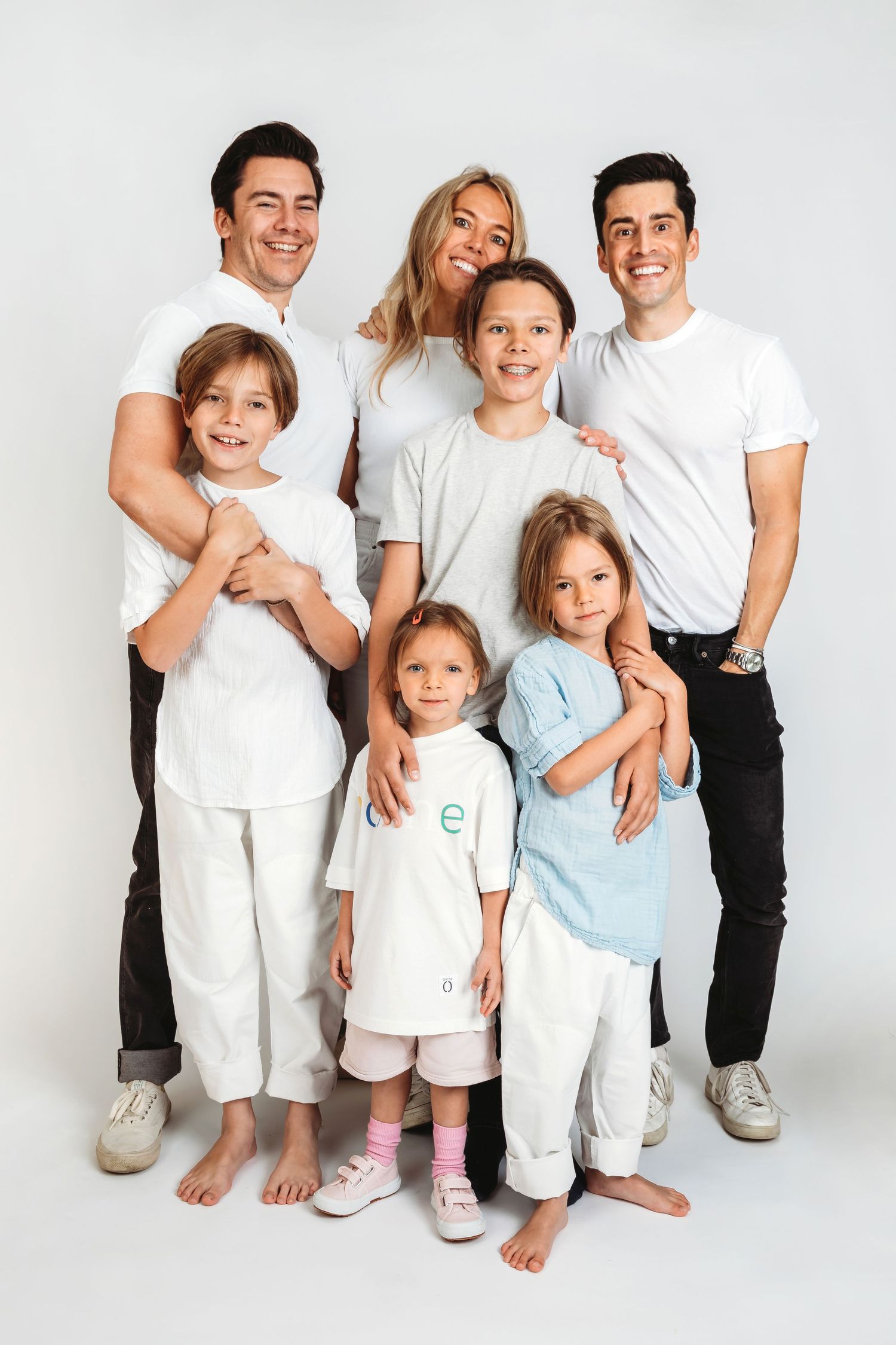 Family of six wearing coordinated white outfits pose together against studio background.