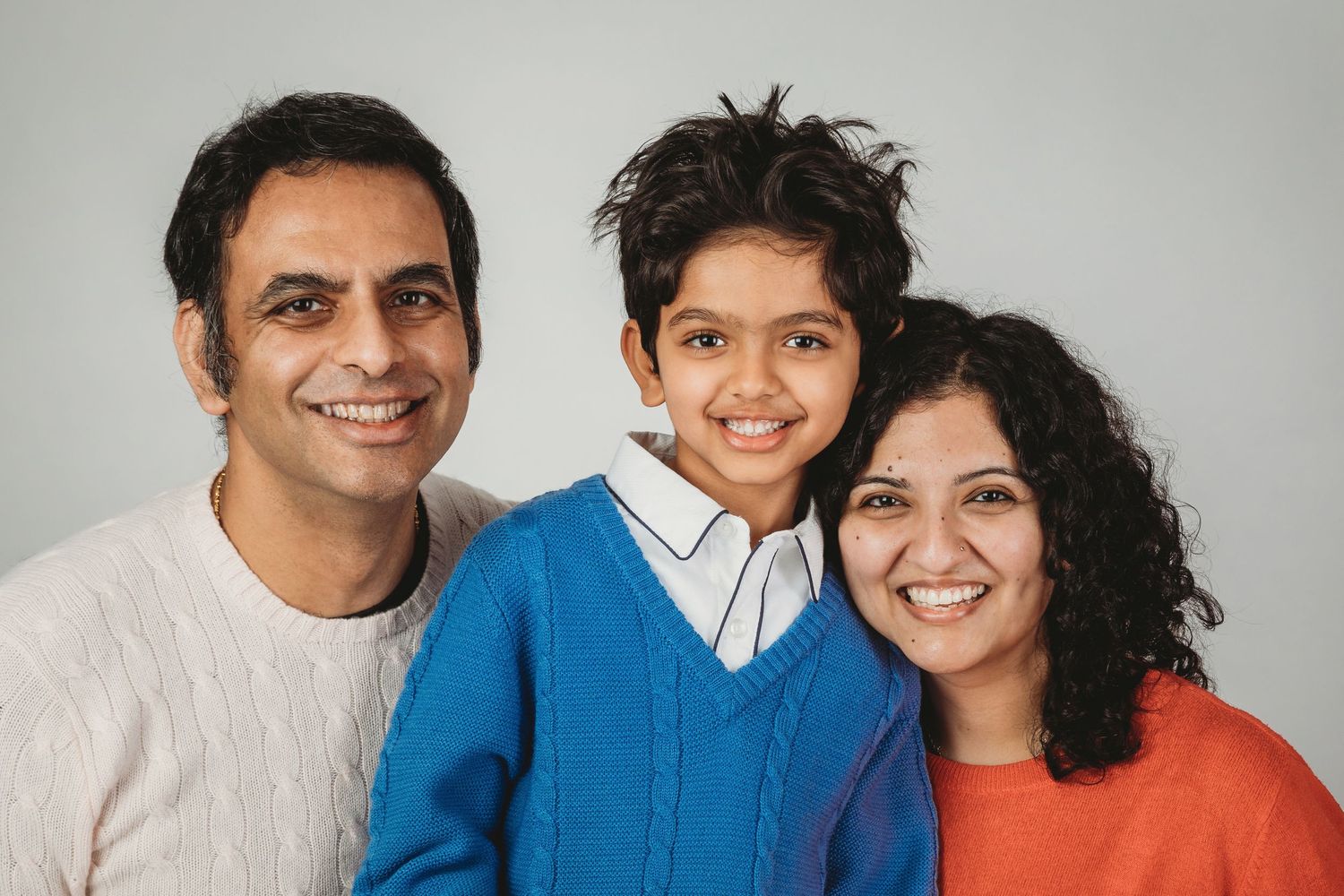A happy family of three poses together against a gray background wearing casual sweaters.