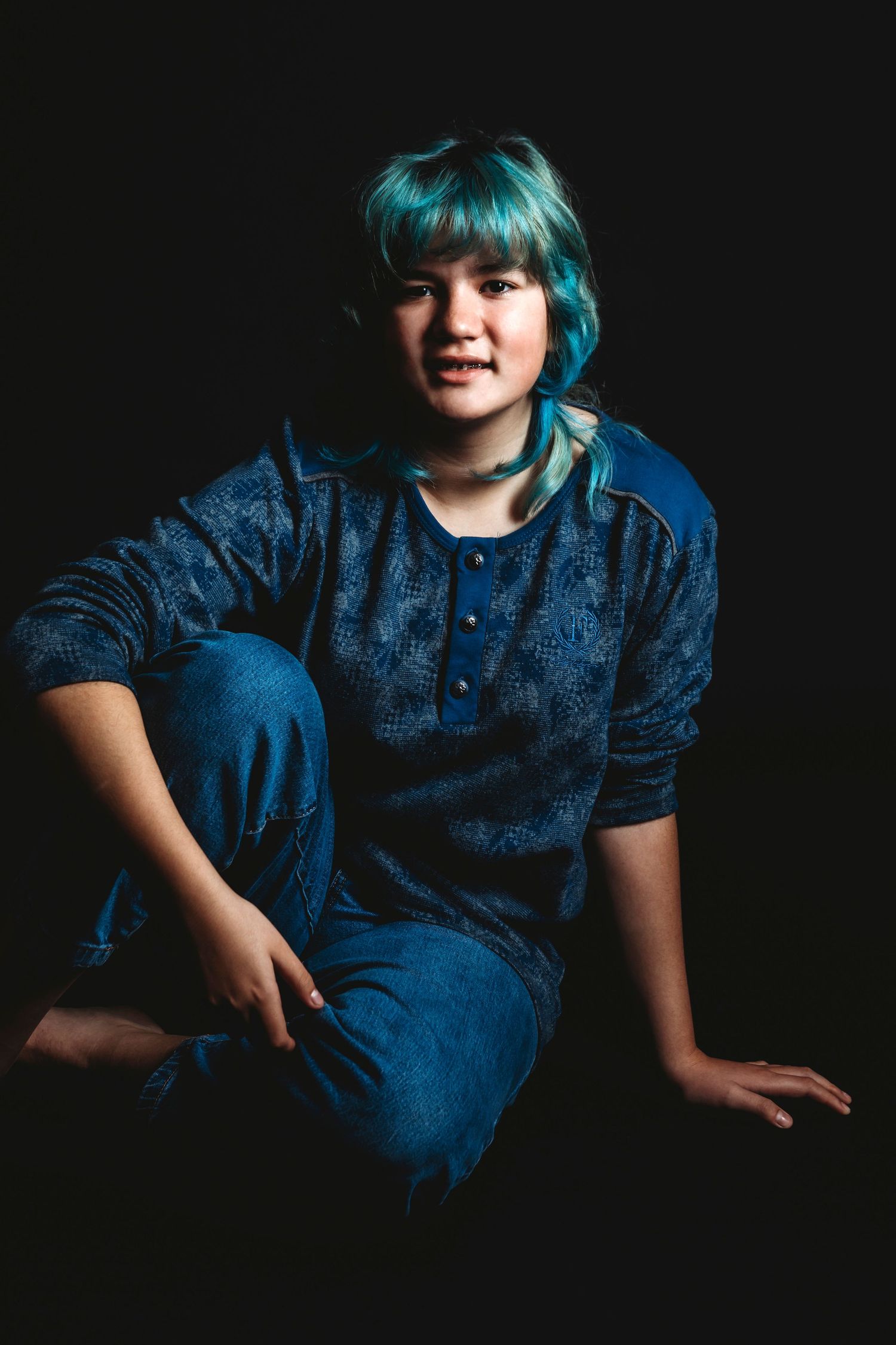 Casual portrait of person with teal hair sitting against dark background.