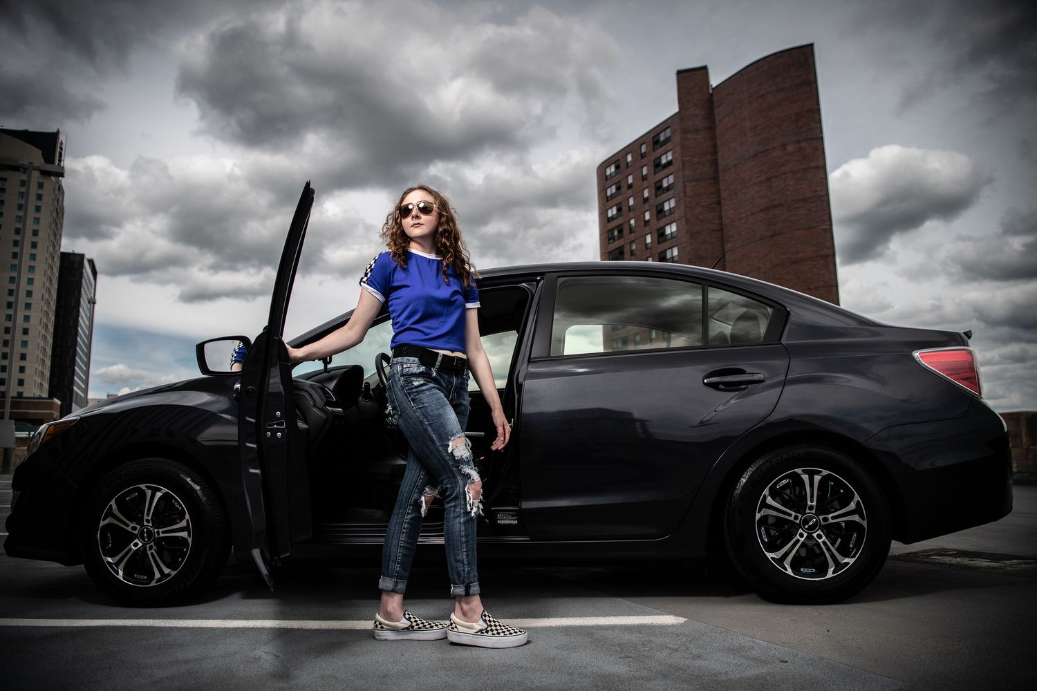 Dramatic photo of someone posed with a black car under stormy skies in an urban setting.