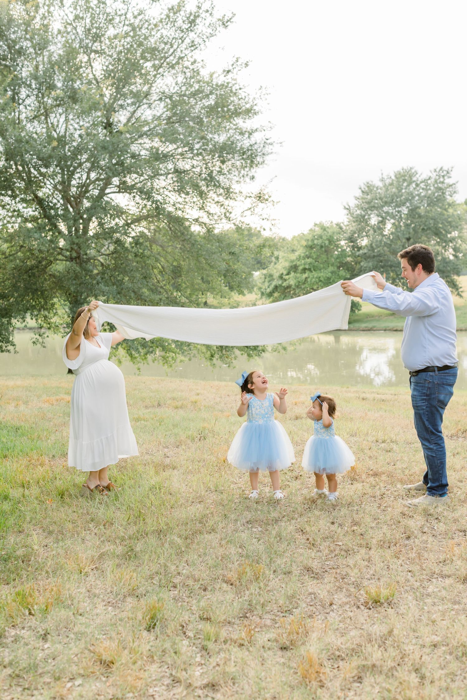 Family holding up white fabric together in grassy field with pond in background.