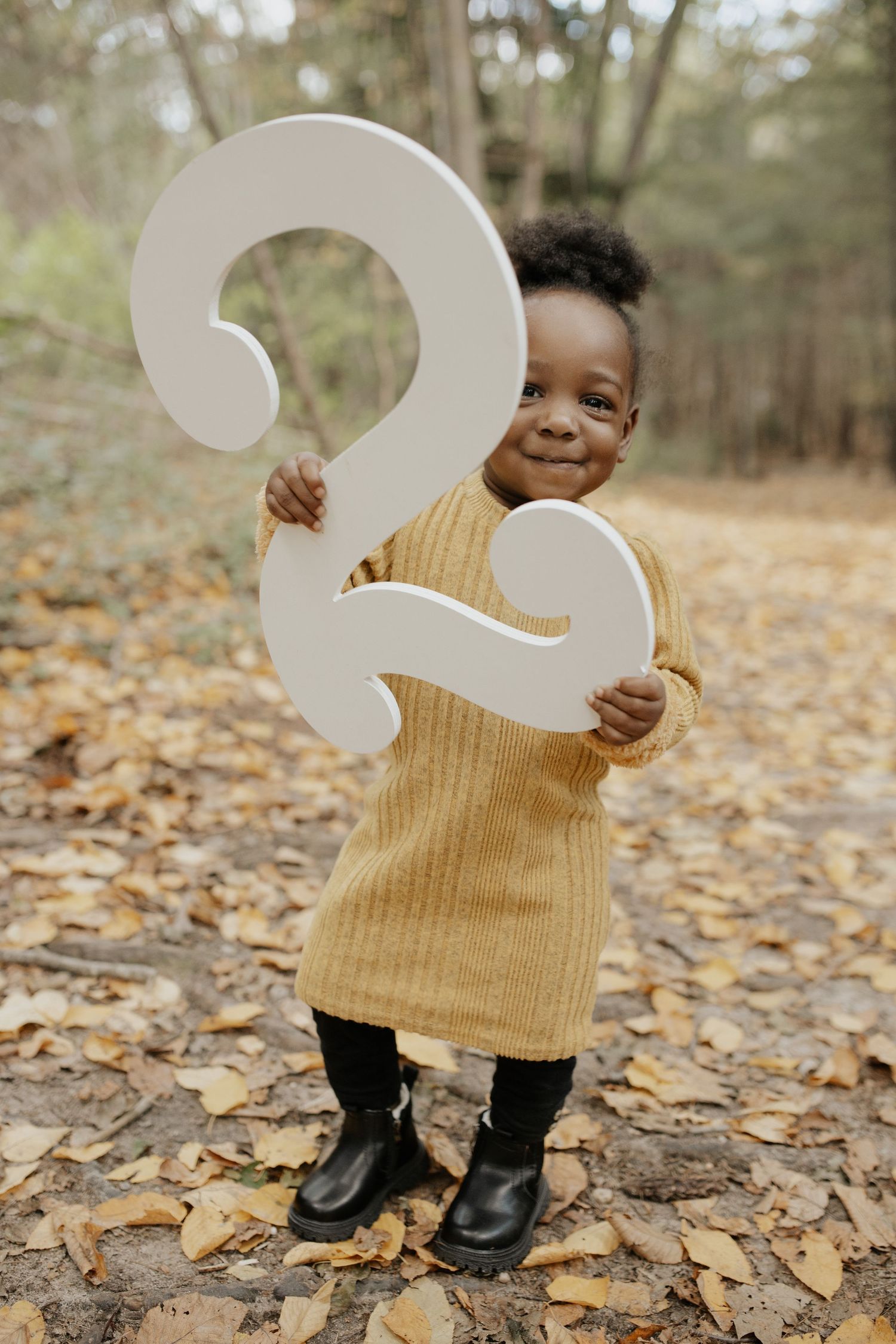 Toddler in yellow sweater dress and boots holding large number 2 prop on autumn ground.