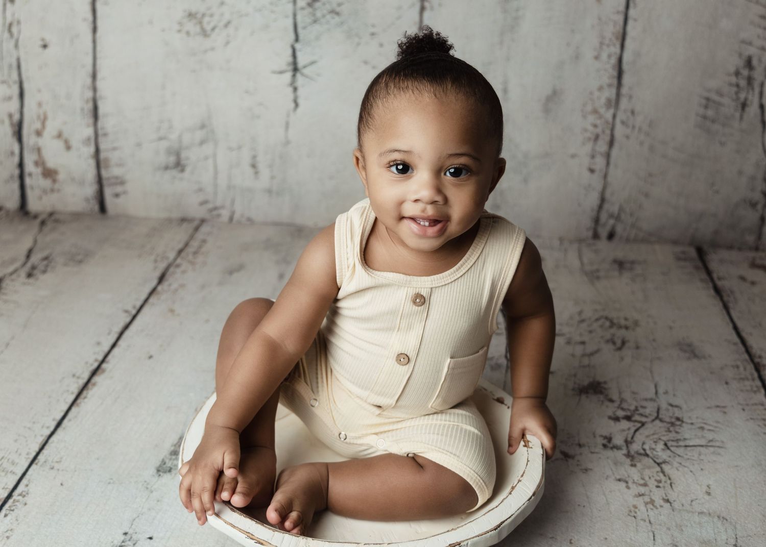 Baby in cream romper sitting on white wooden floor with bright expression.