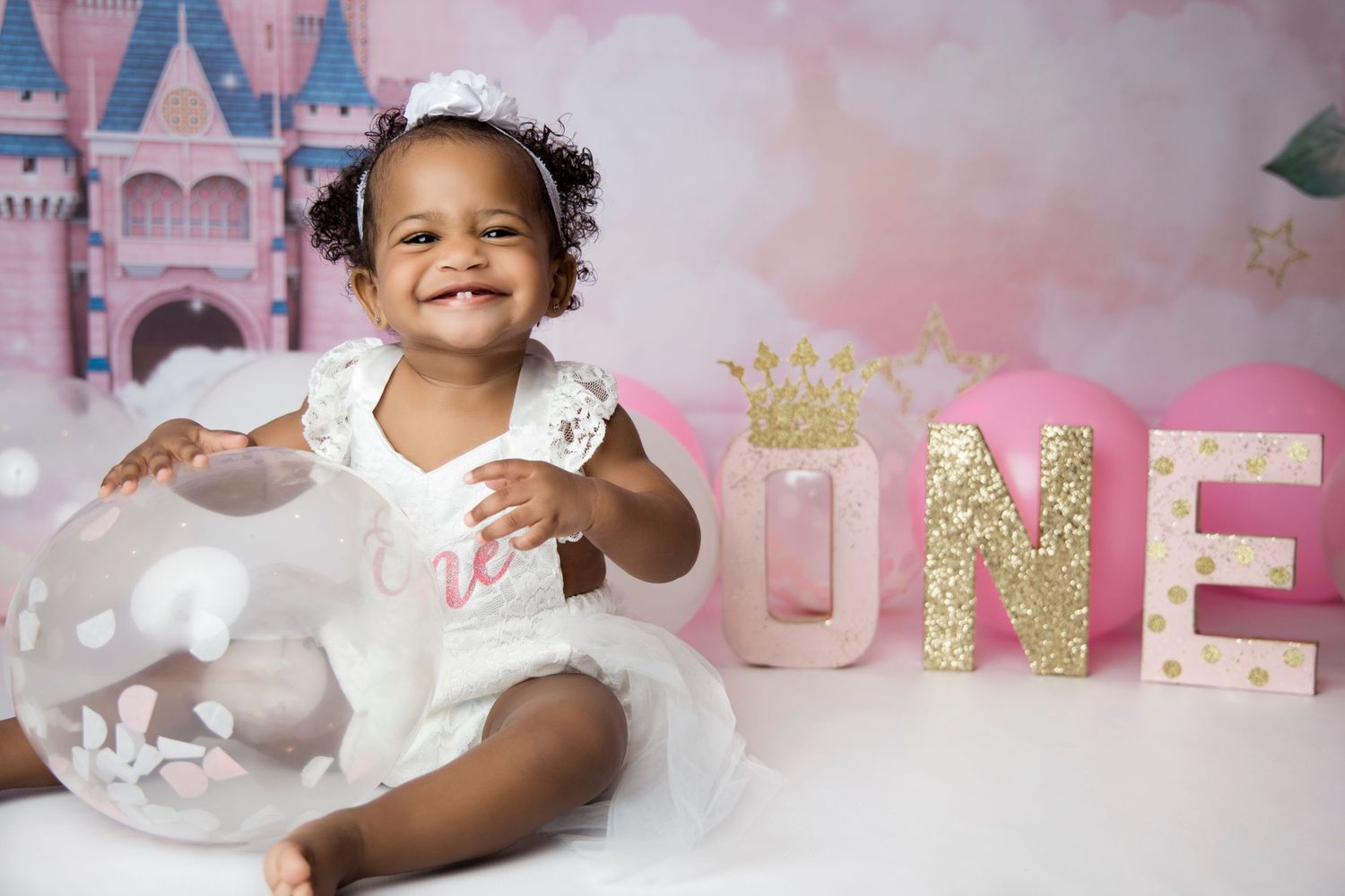 Baby in white dress celebrates first birthday with castle backdrop and gold letters.