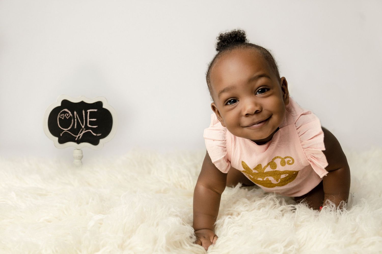 Baby in pink ruffled outfit crawling on white rug with 'ONE' sign.