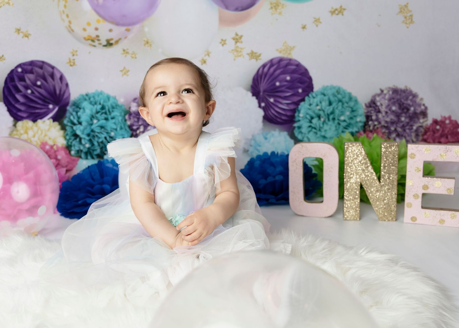 Baby in white dress at first birthday celebration with purple and teal decorations.