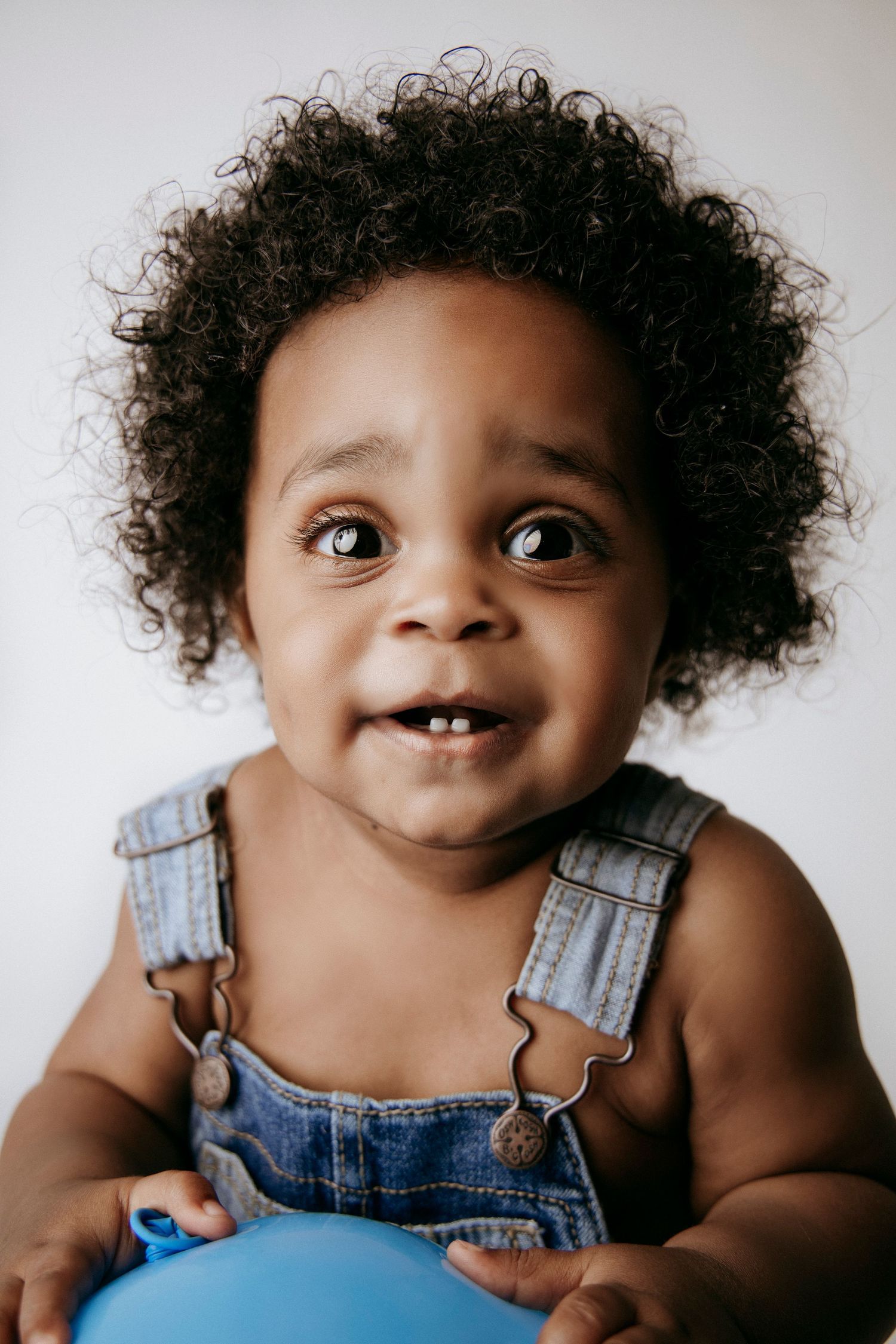 Joyful toddler in denim overalls smiling at camera with natural curly hair.