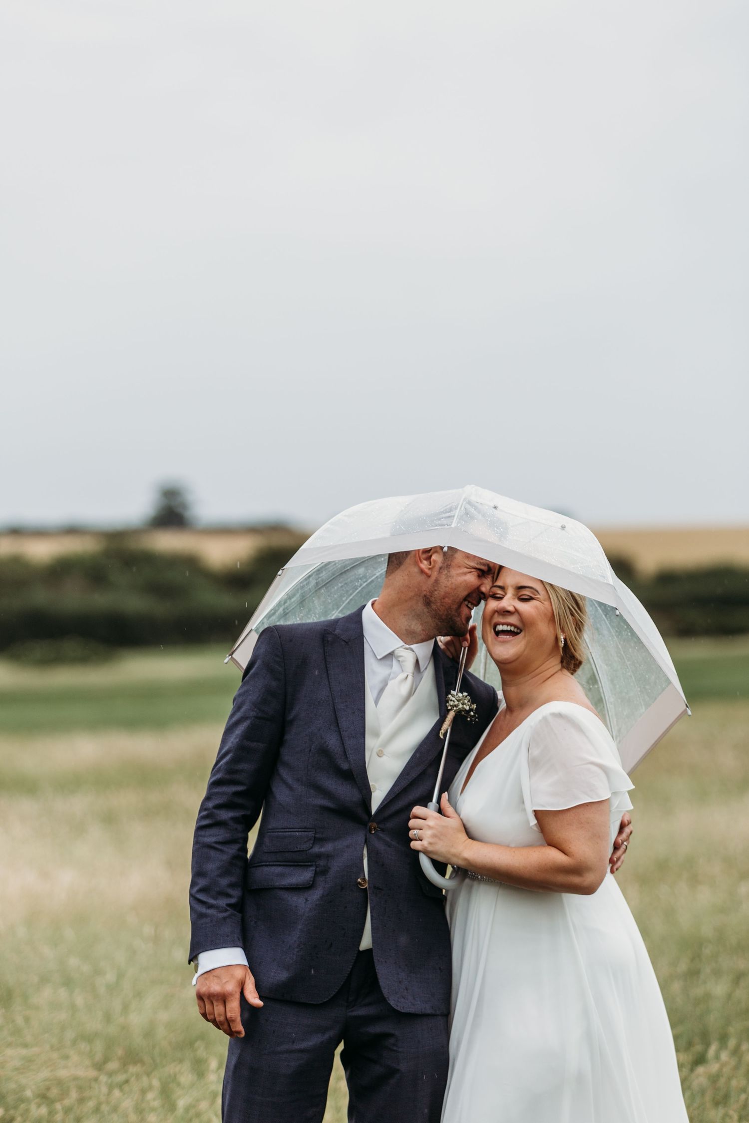 A romantic wedding couple shares a laugh under a veil in an outdoor field setting.