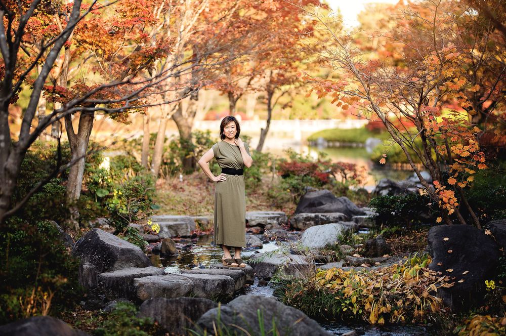 Person in elegant dress poses among autumn foliage and rocks near a stream.
