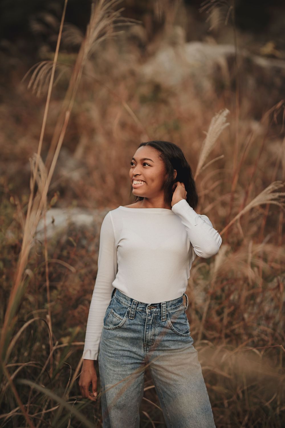 Person in white sweater and jeans poses naturally among tall autumn grasses during golden hour.