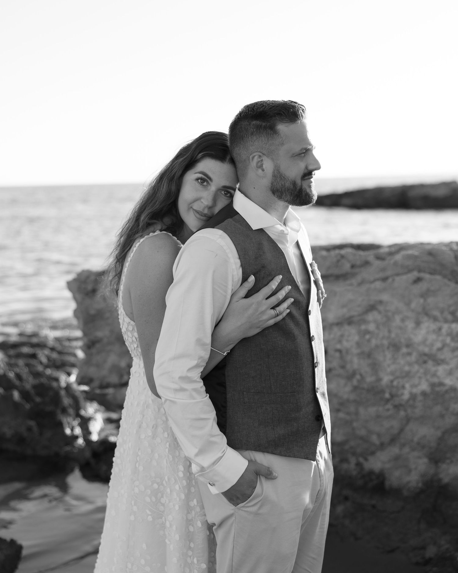 Romantic black and white portrait of newlyweds embracing on rocky coastline at sunset in Lindos, Greece.