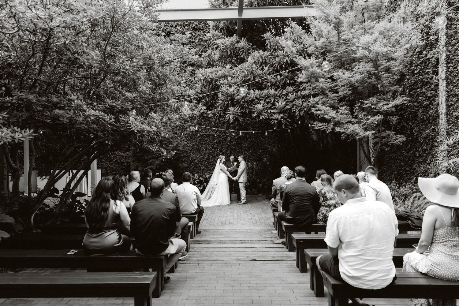 Black and white photo of an outdoor woodland wedding ceremony with guests seated on wooden benches.