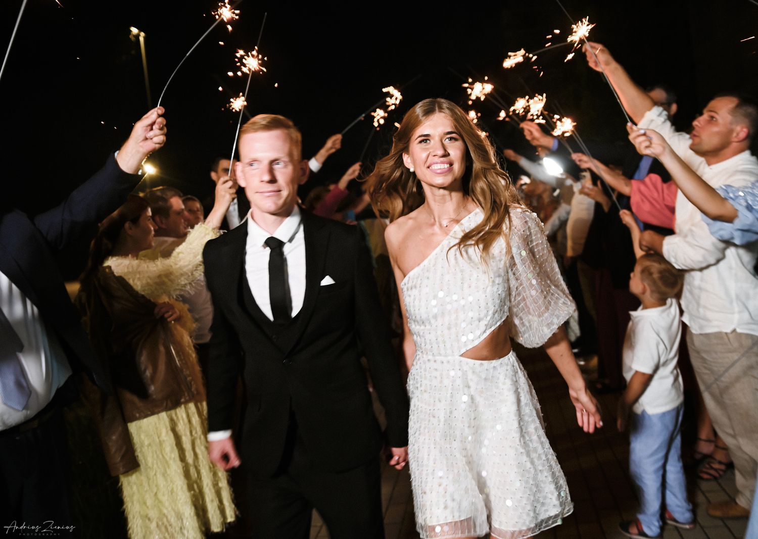 Wedding couple celebrating with sparklers surrounded by guests during evening reception.